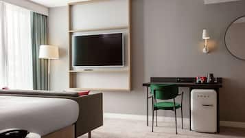 A hotel room with a bed, a desk with a chair, a television, a refrigerator, and a lamp.