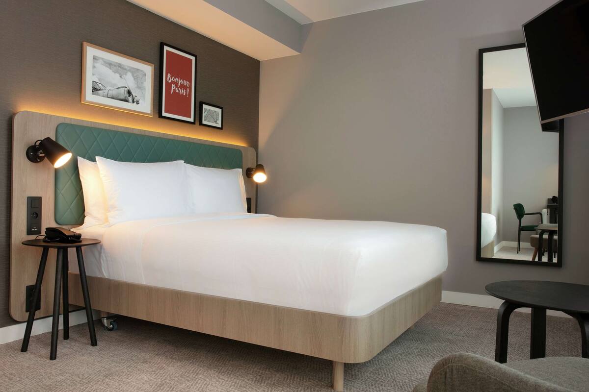 A modern hotel room with a large bed, a bedside table with a lamp, a small table, and a mirror on the wall.