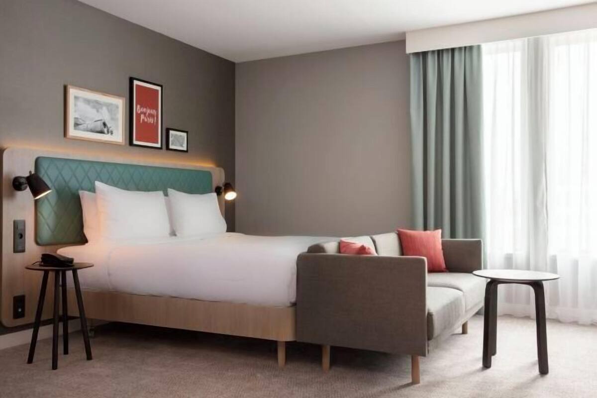 A hotel room with a bed, a sofa, a small table, and a nightstand. There are framed pictures on the wall and a window with curtains.