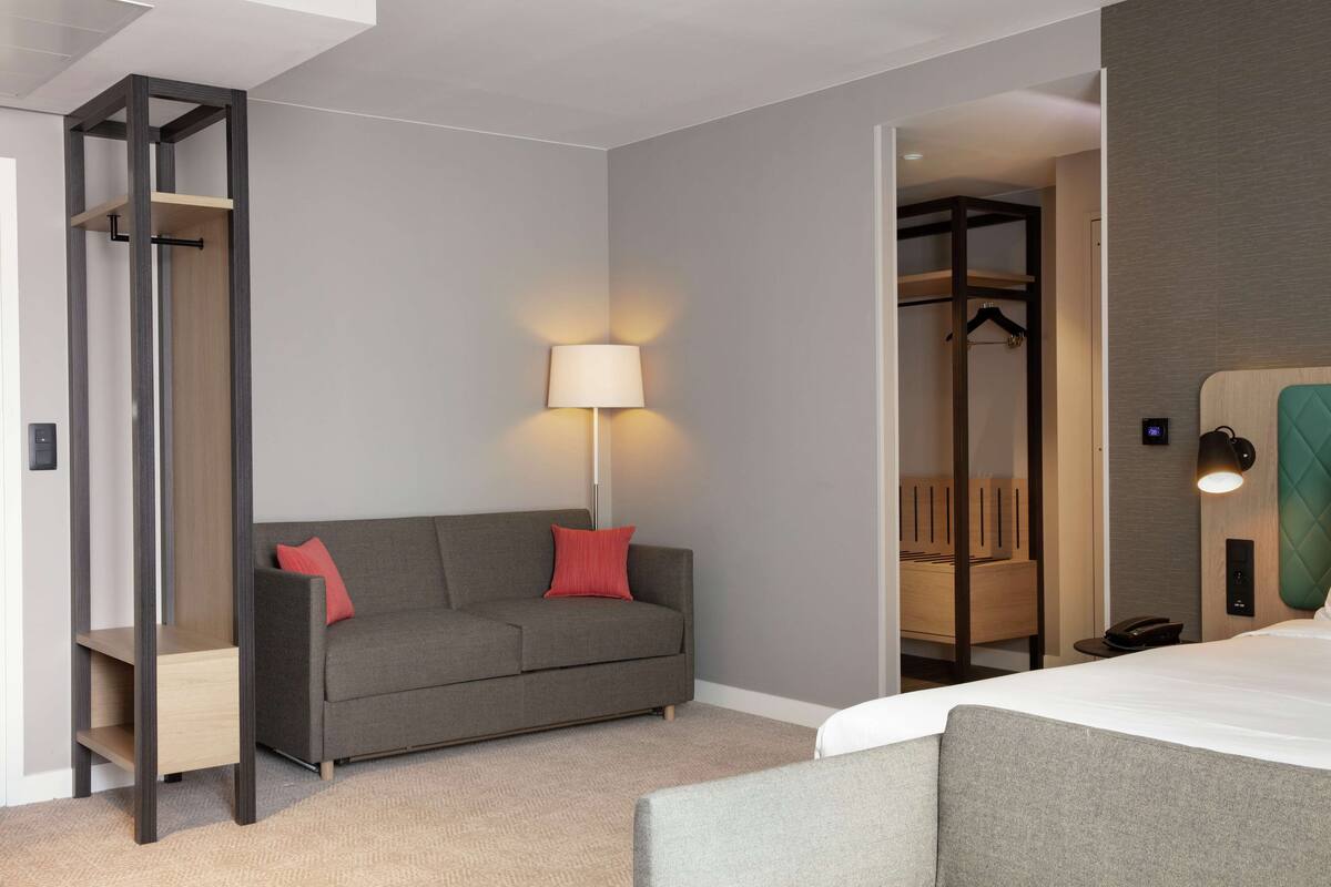 A hotel room with a grey sofa, a bed, a lamp, and a walk-in closet.