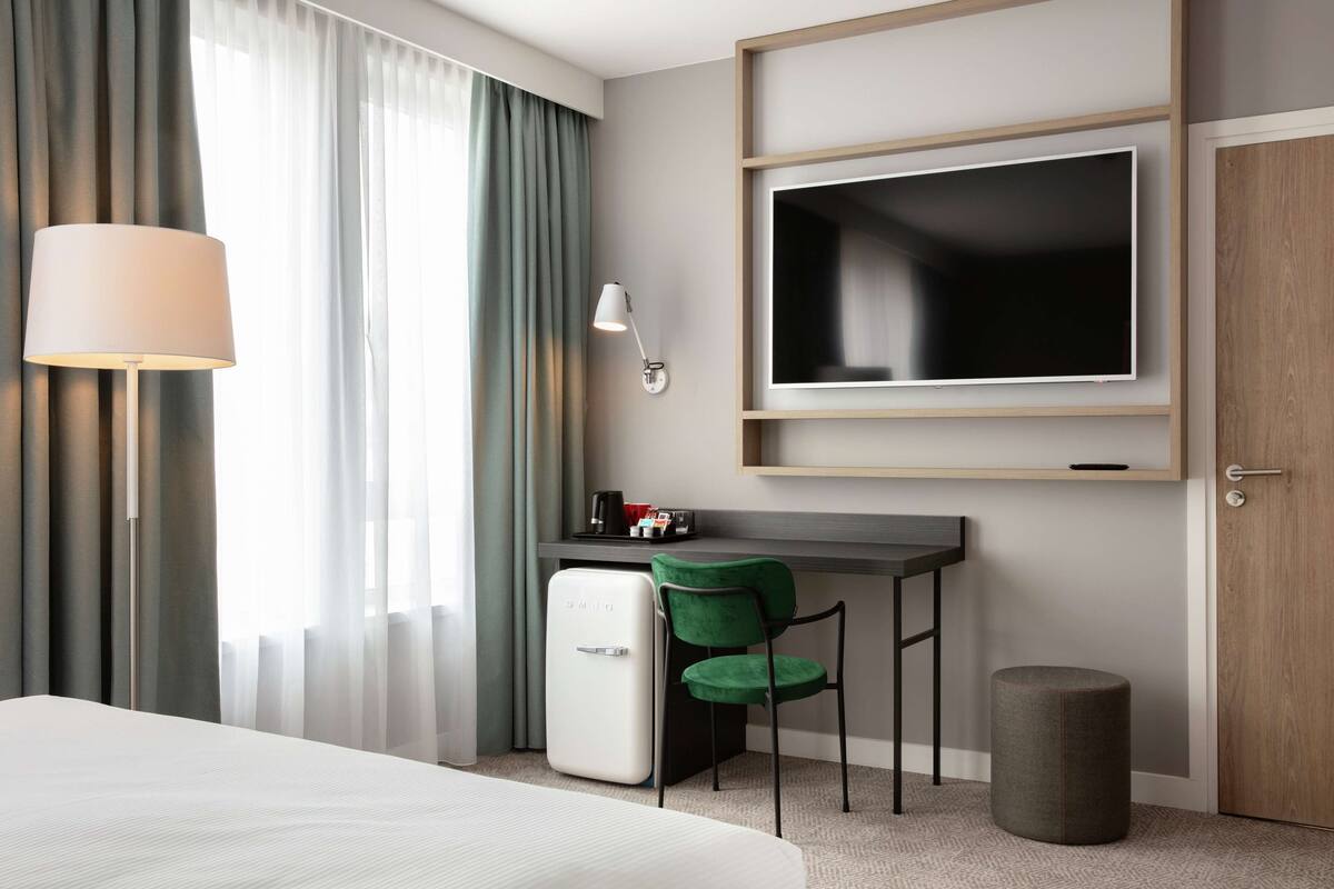 A hotel room with a bed, a desk with a TV, a mini-fridge, a chair, a lamp, and a door.