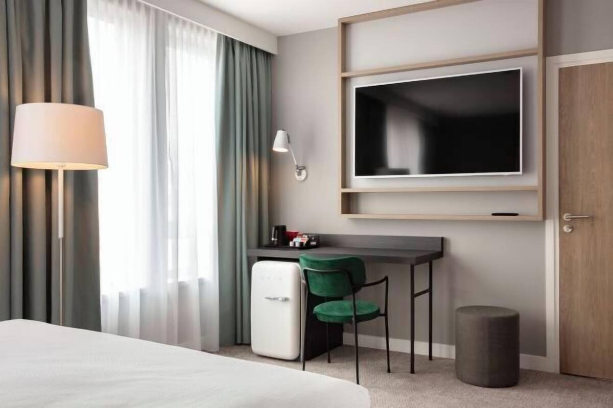 A hotel room with a bed, a desk with a chair, a television, a lamp, and a small trash bin.