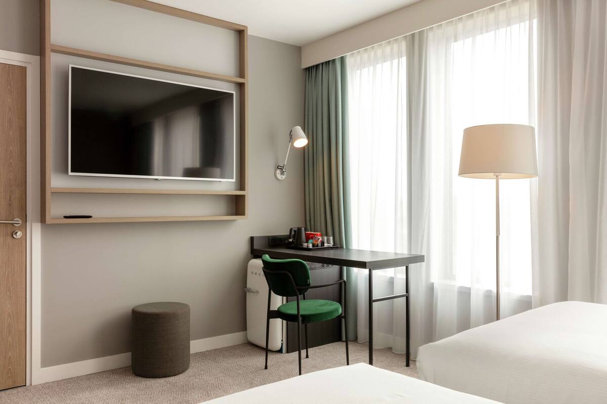 A hotel room with a bed, a desk with a chair, a TV mounted on the wall, a lamp, and a window with curtains.