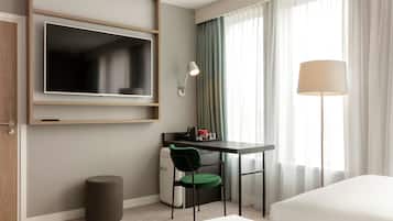 Una habitación de hotel con cama, escritorio con silla, televisor montado en la pared, lámpara y ventana con cortinas.