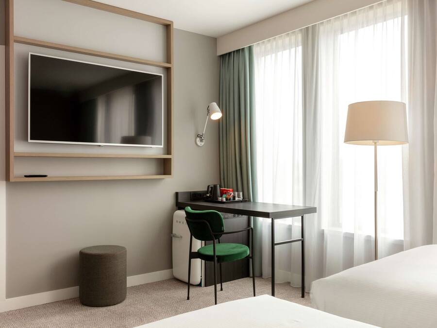 Una habitación de hotel con cama, escritorio con silla, televisor montado en la pared, lámpara y ventana con cortinas.