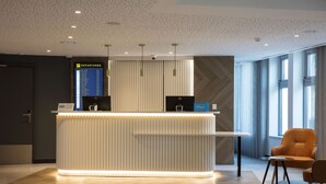 Un moderno mostrador de recepción de hotel con un panel informativo digital de salidas, una zona de estar con una silla y una mesita, y un amplio ventanal que proporciona luz natural.