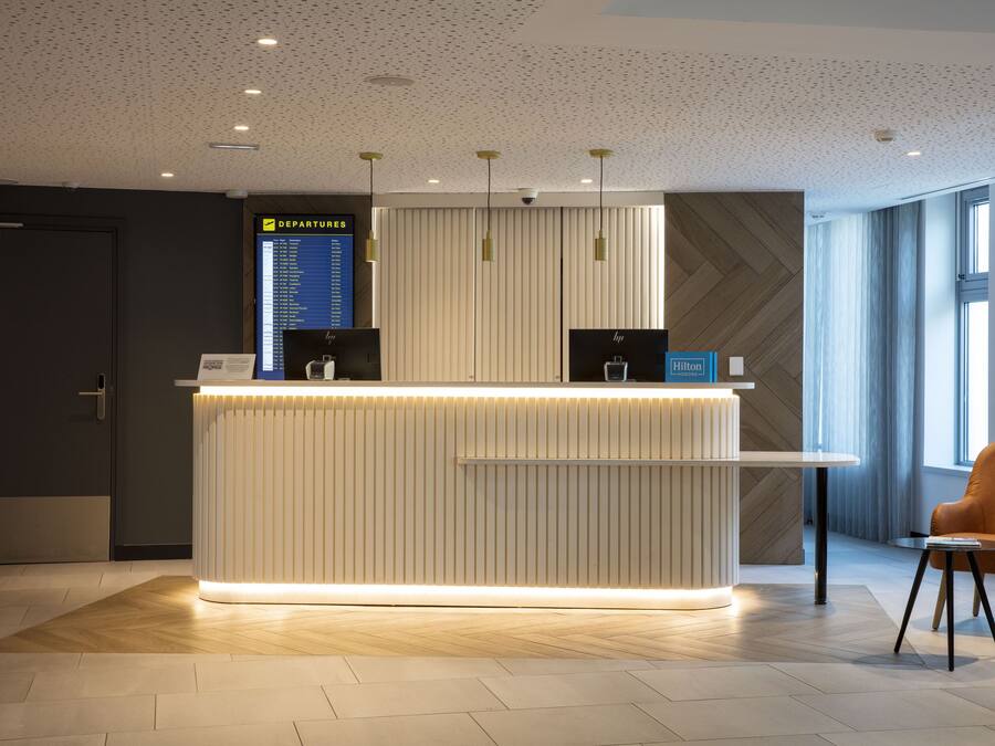 A modern hotel reception desk with a digital departures board, a seating area with a chair and a small table, and a large window providing natural light.