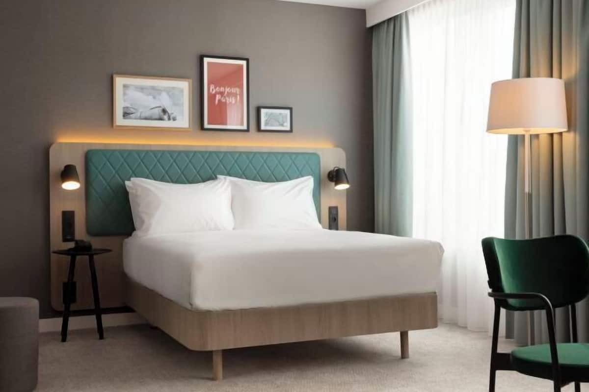A hotel room with a bed, two bedside tables, a green chair, a lamp, and framed pictures on the wall.