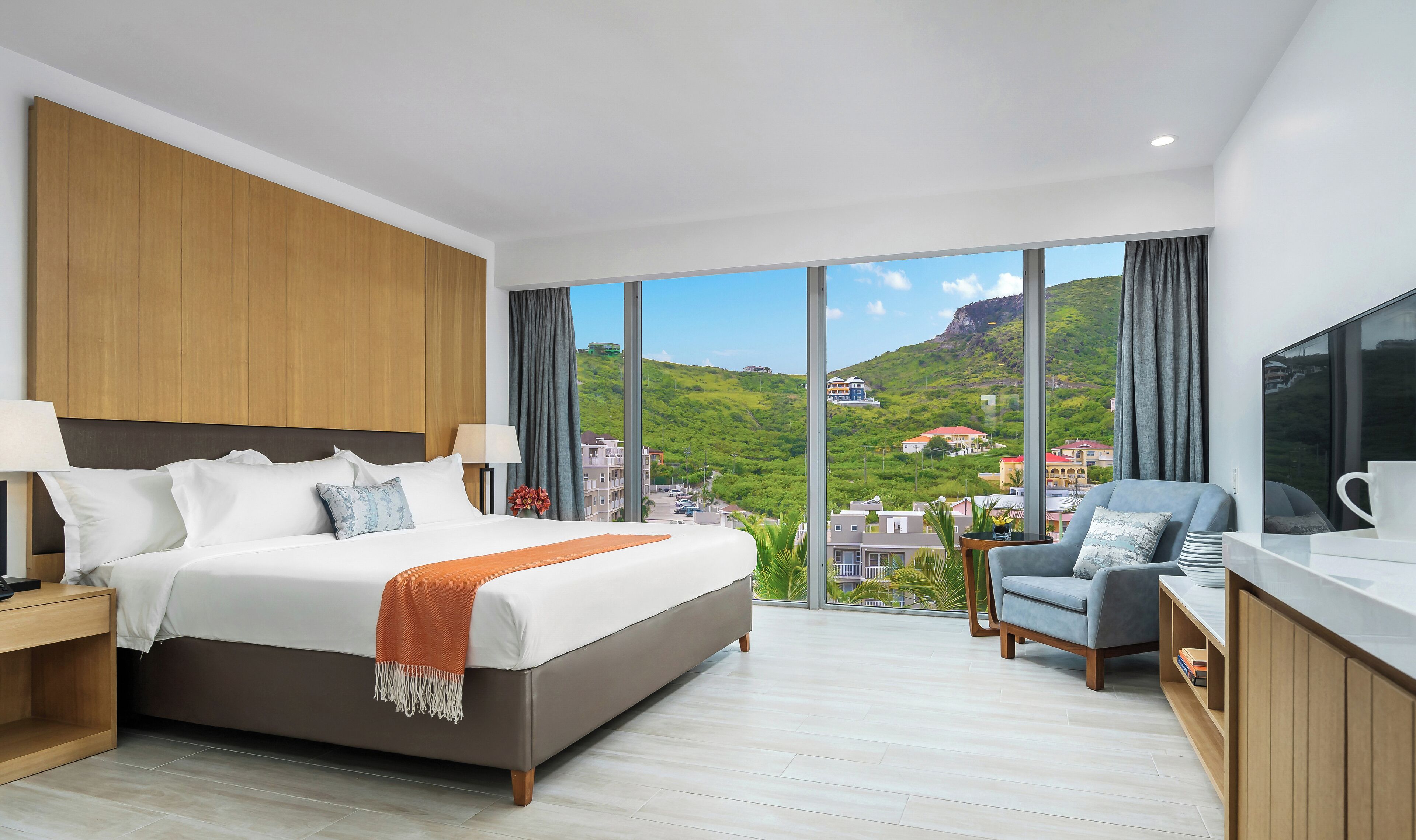 A modern hotel room with a large bed, a desk, and a view of a green hillside.