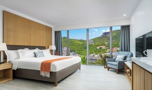 A modern hotel room with a large bed, a desk, and a view of a green hillside. - Koi Resort Saint Kitts, Curio Collection by Hilton (Basseterre)