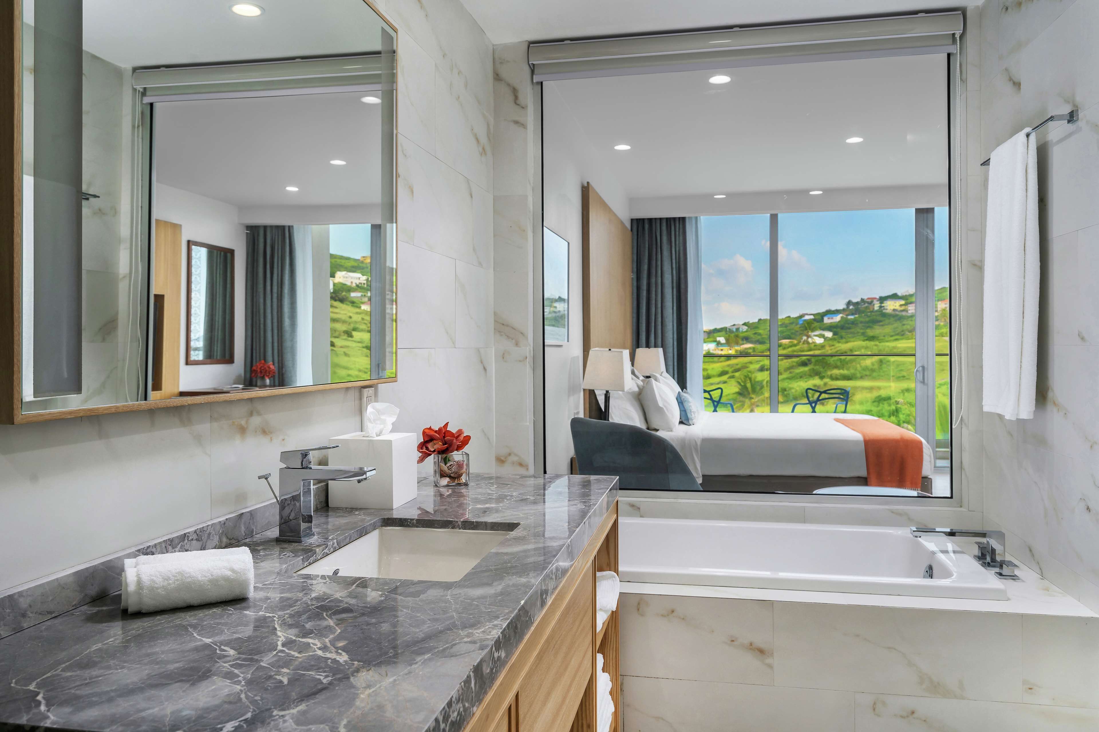 A modern bathroom with a large mirror, a marble sink, a bathtub, and a view of a green landscape through large windows.
