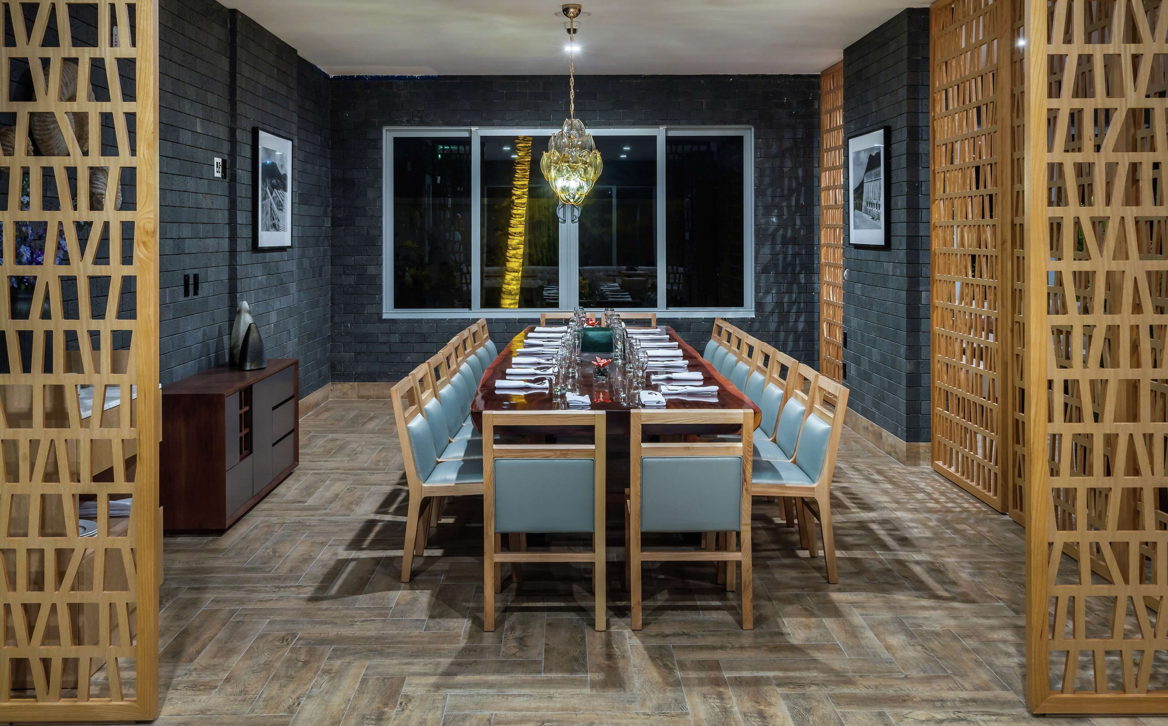 A dining area with a long wooden table set for six, flanked by wooden chairs with light blue cushions. The room features dark brick walls, large windows, and a hanging light fixture.