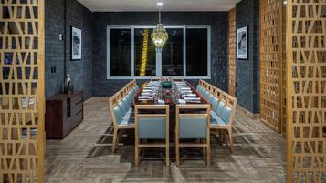 A dining area with a long wooden table set for six, flanked by wooden chairs with light blue cushions. The room features dark brick walls, large windows, and a hanging light fixture.