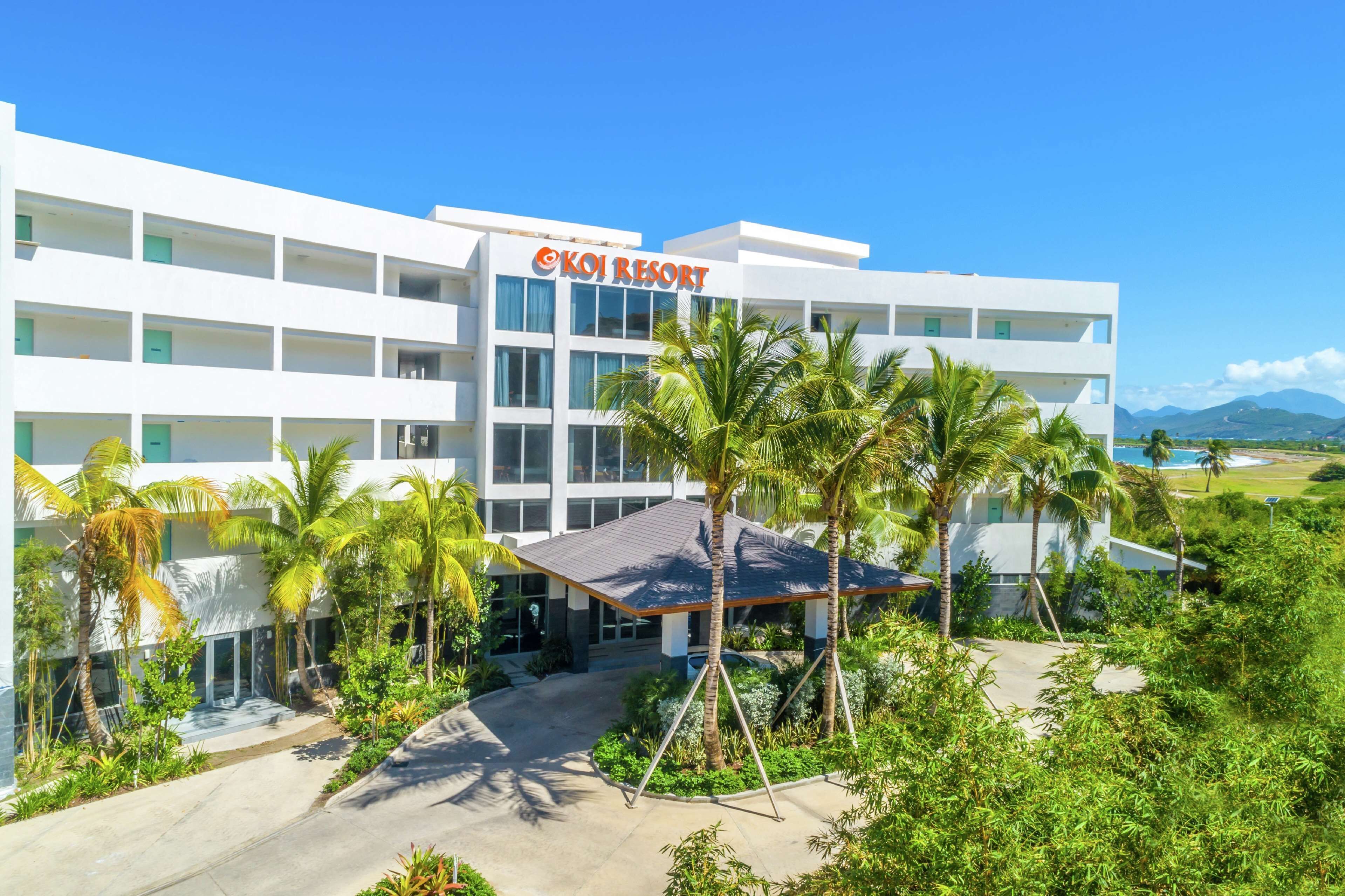 Un hotel moderno con un letrero llamativo que dice "Koi Resort". Cuenta con una estructura de varios pisos con balcones, rodeada de exuberante follaje y palmeras.