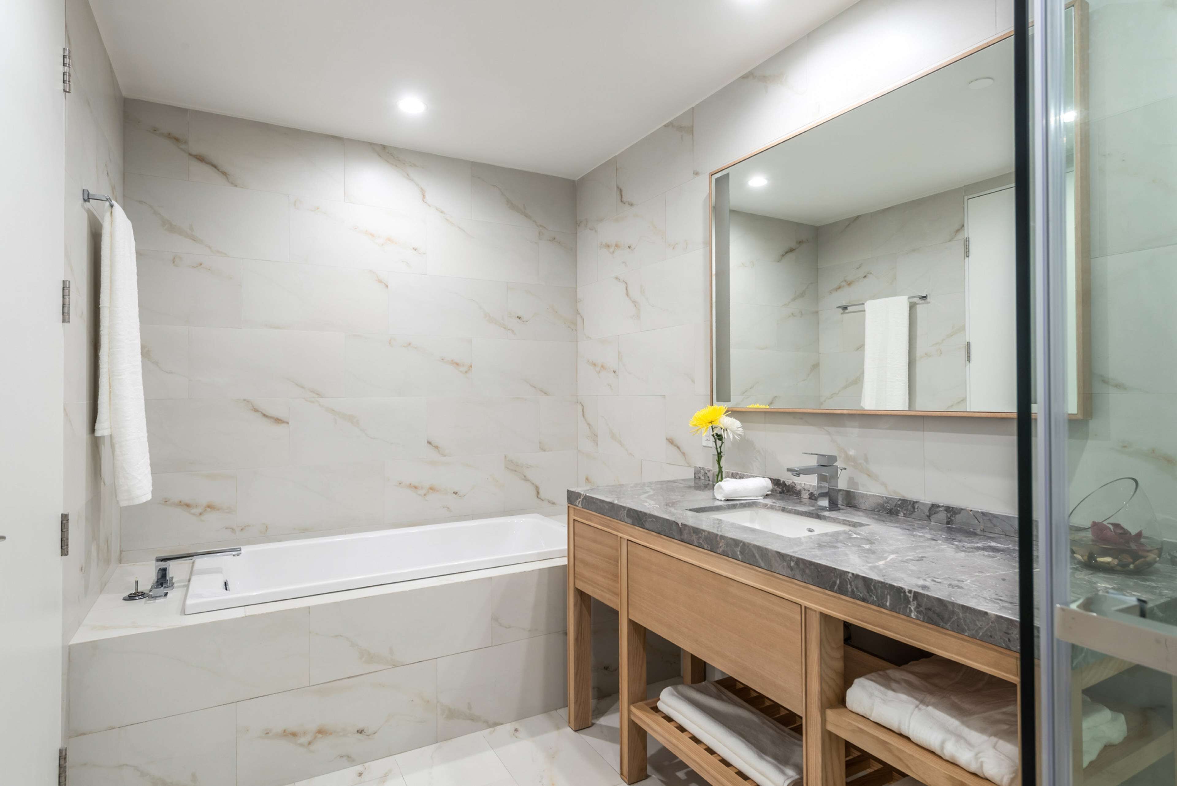 A modern bathroom with a marble wall, a bathtub, a sink with a mirror, and wooden shelves.