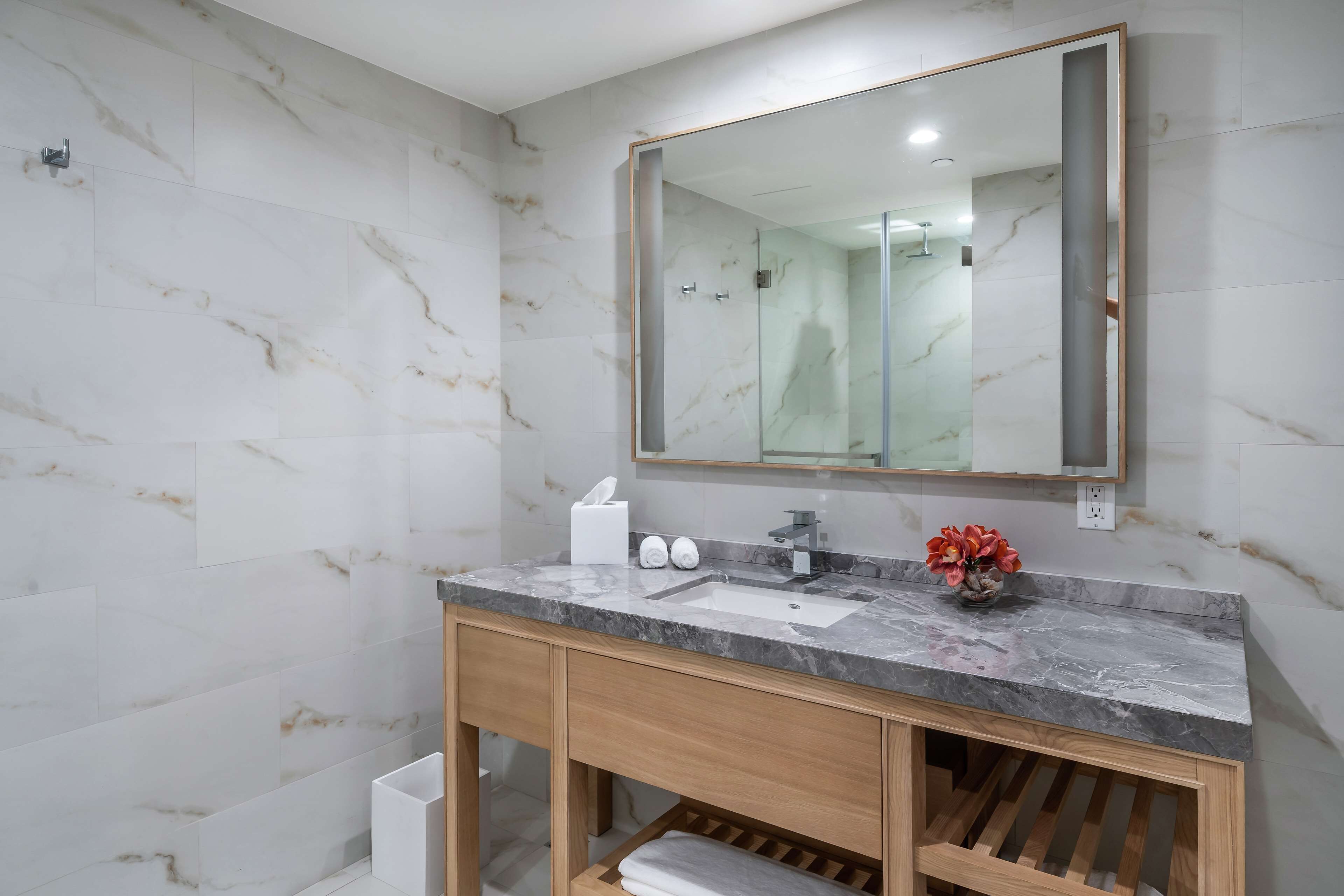 A modern bathroom with a marble-tiled wall, a large mirror, a wooden vanity, and a glass shower enclosure.