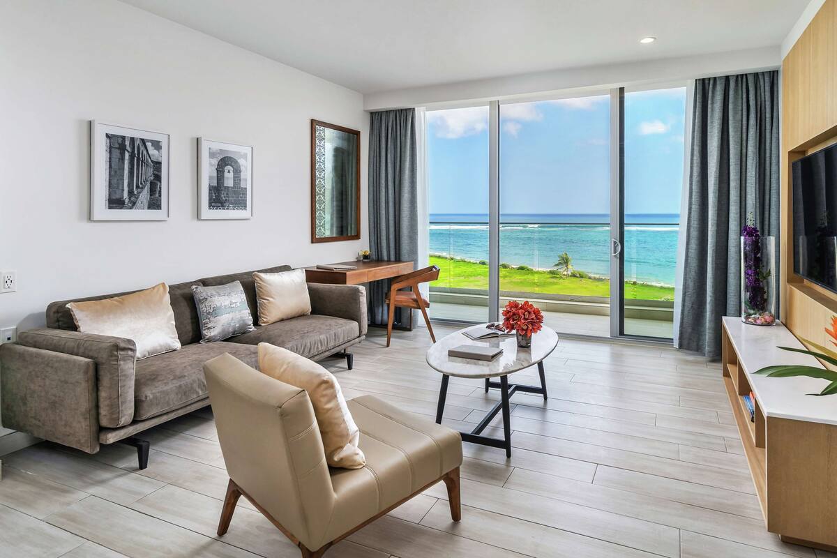 A modern living room with a sofa, armchair, coffee table, and a view of the ocean through large sliding glass doors.