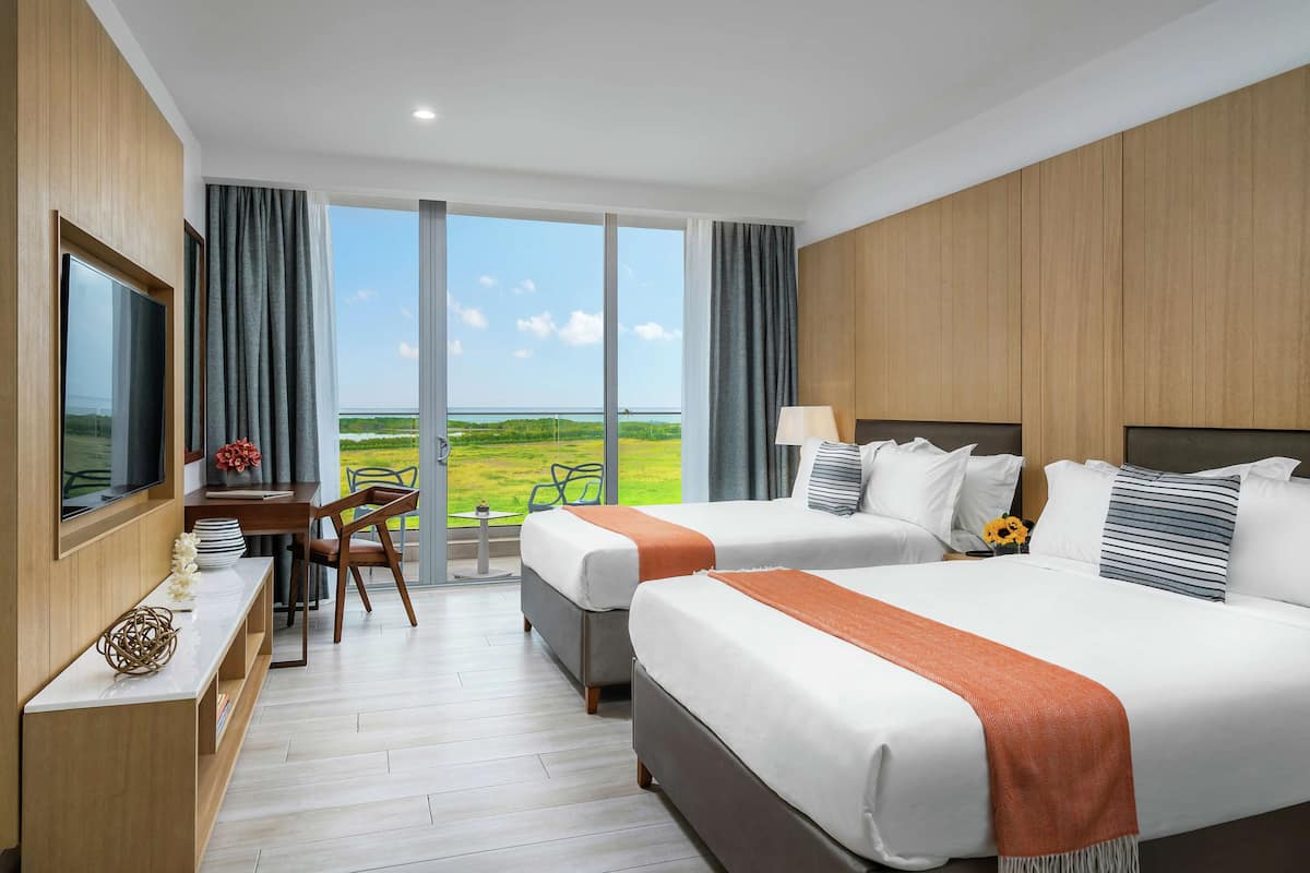 A hotel room with two beds, a desk, and a large window offering a view of the landscape.