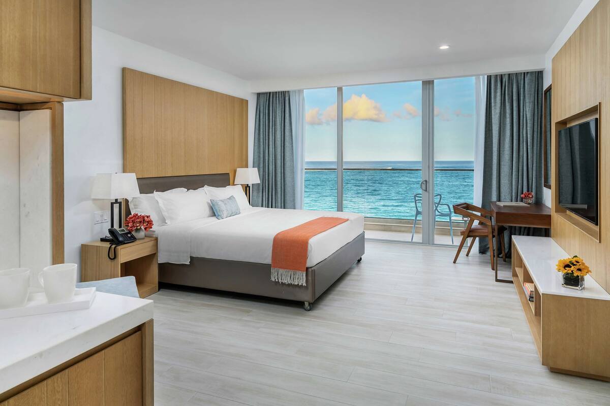 A modern hotel room with a large bed, a desk, a chair, and a TV. The room has a view of the sea through large windows.