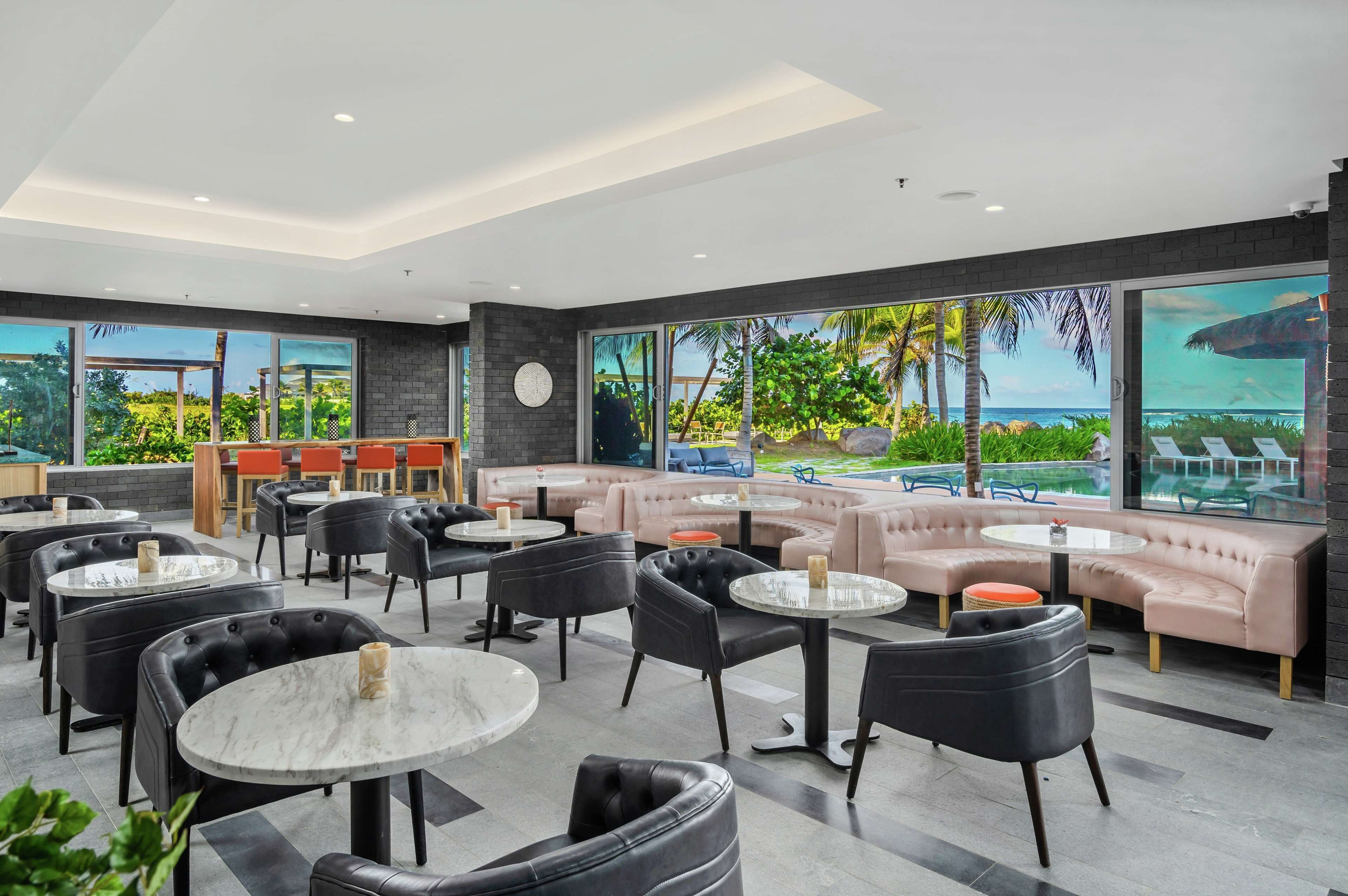 Un espace de détente moderne avec des canapés en cuir, des tables rondes et une grande fenêtre donnant sur un jardin tropical et l’océan.