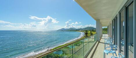 A balcony with a view of the ocean and beach, featuring glass railings and blue chairs.