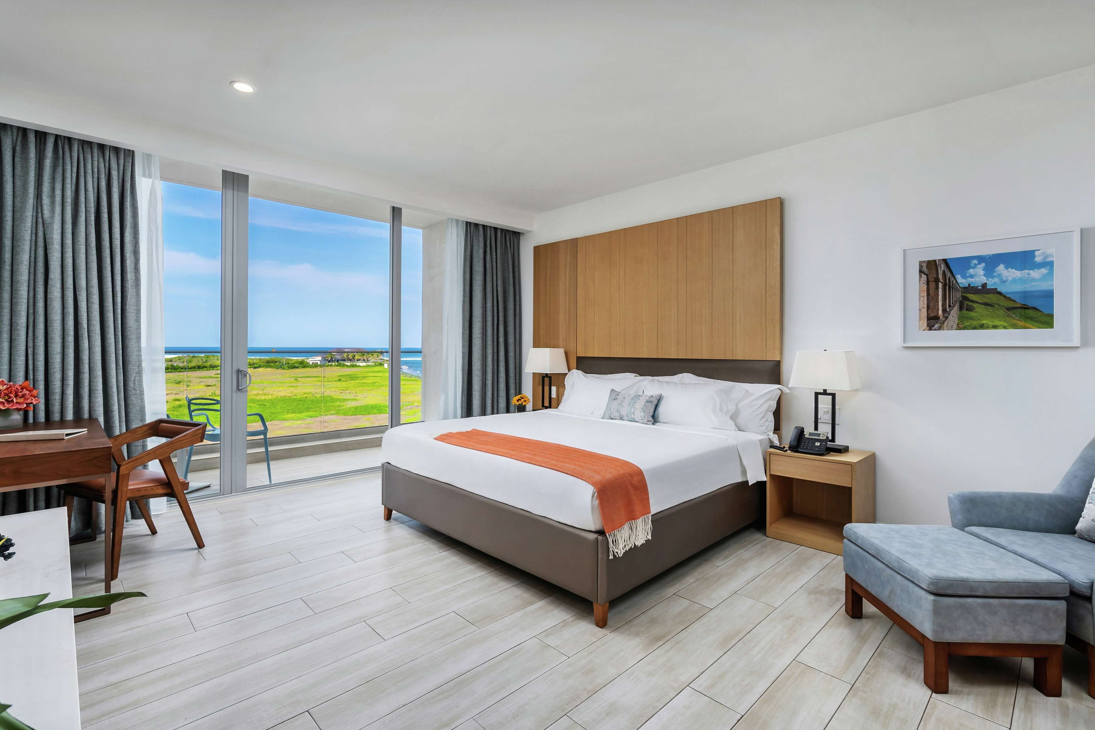 A modern hotel room with a large bed, a desk with a chair, a sofa, and a view of the sea.
