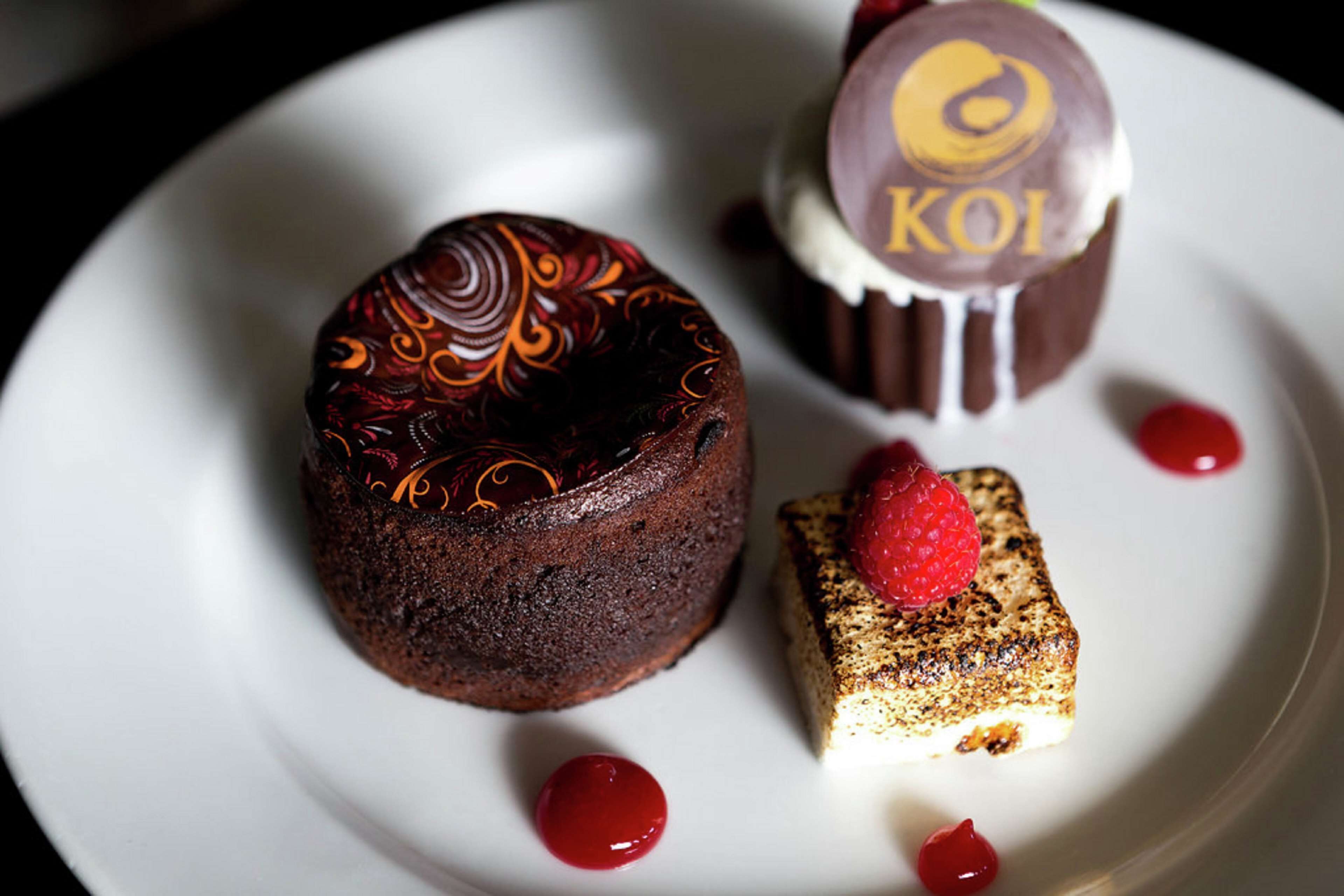 A dessert plate with a chocolate cake, a chocolate-covered treat, and a small square cake with a raspberry on top.