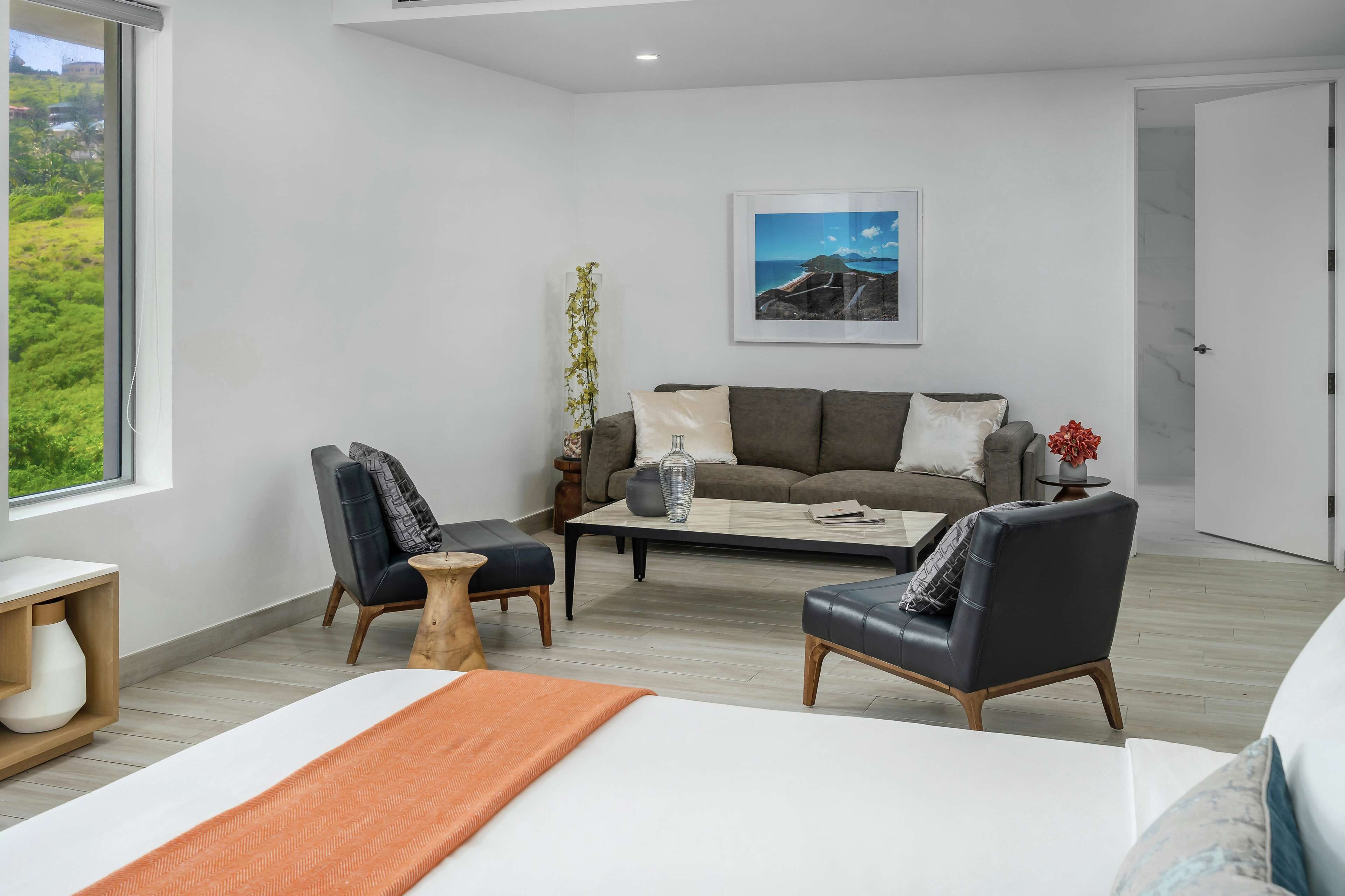 A modern hotel room with a bed, two armchairs, a sofa, a coffee table, and a large window offering a view of greenery.