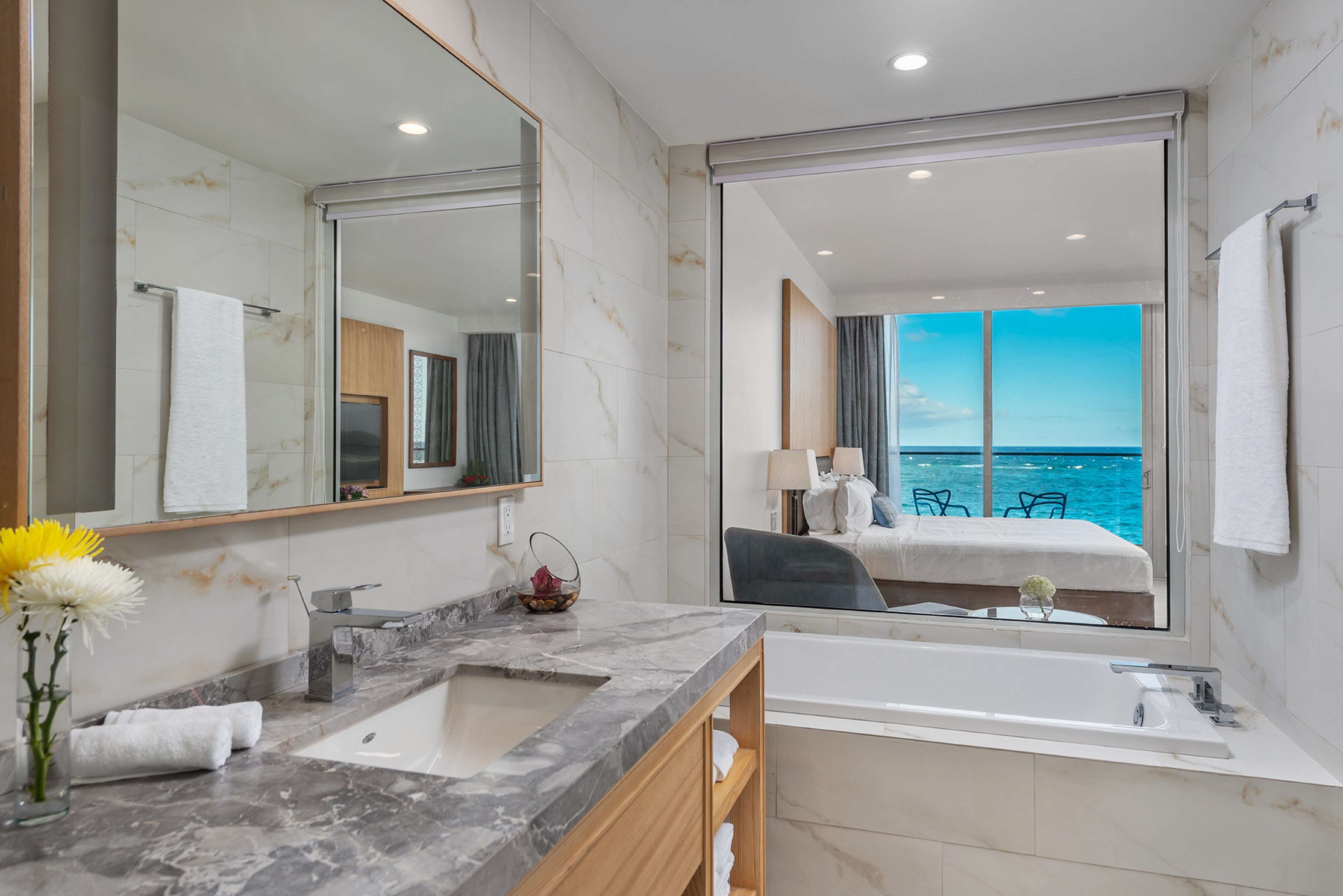 A modern bathroom with a large mirror, marble countertop, and a bathtub. There is a view of a balcony with a chair and a table, and a window overlooking the ocean.