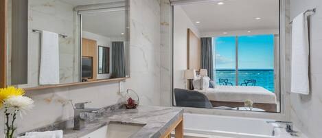A modern bathroom with a large mirror, marble countertop, and a bathtub. There is a view of a balcony with a chair and a table, and a window overlooking the ocean.