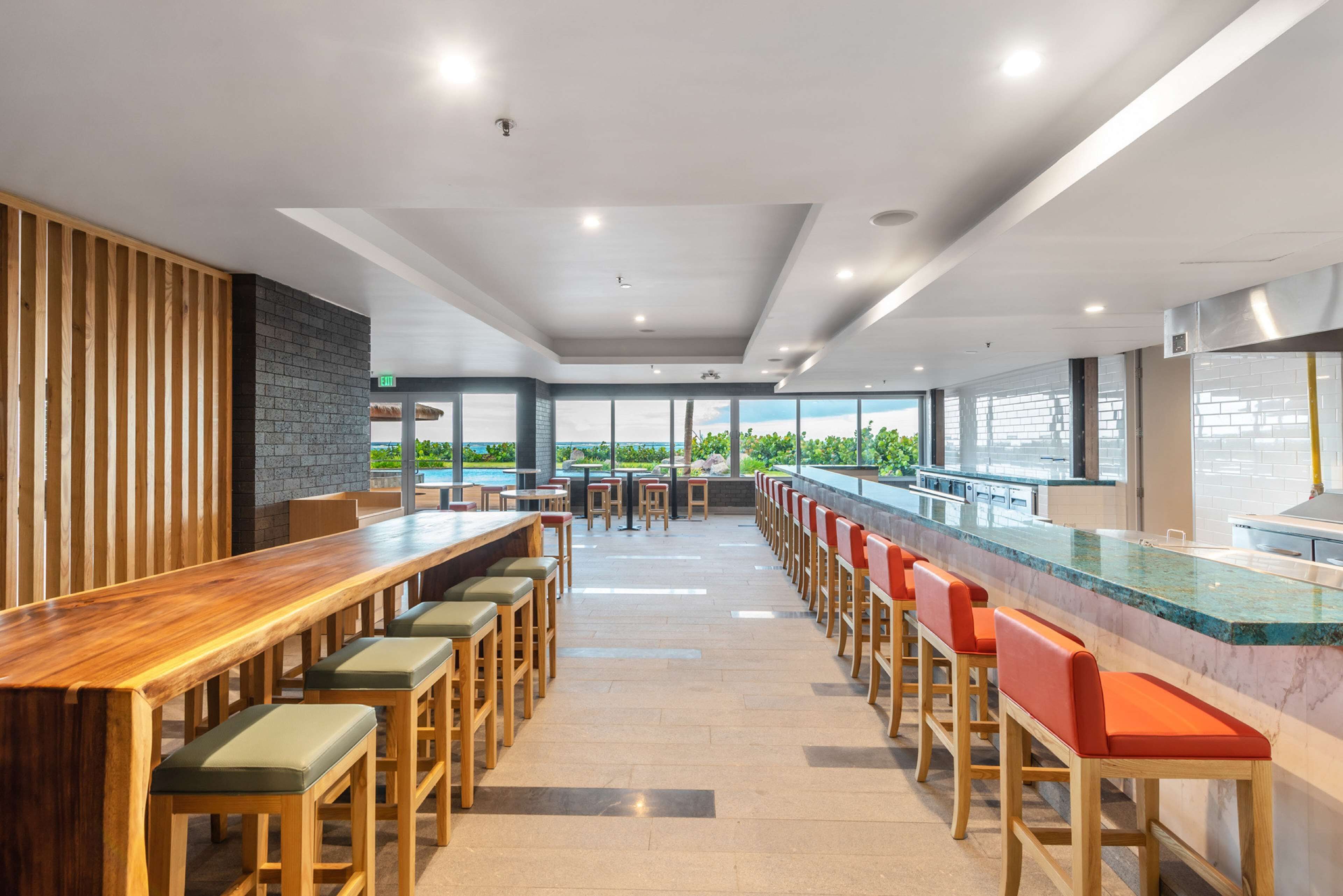 Un restaurant moderne avec de longues tables en bois et des tabourets de bar rouges et confortables, ainsi que de grandes fenêtres donnant sur l’extérieur.
