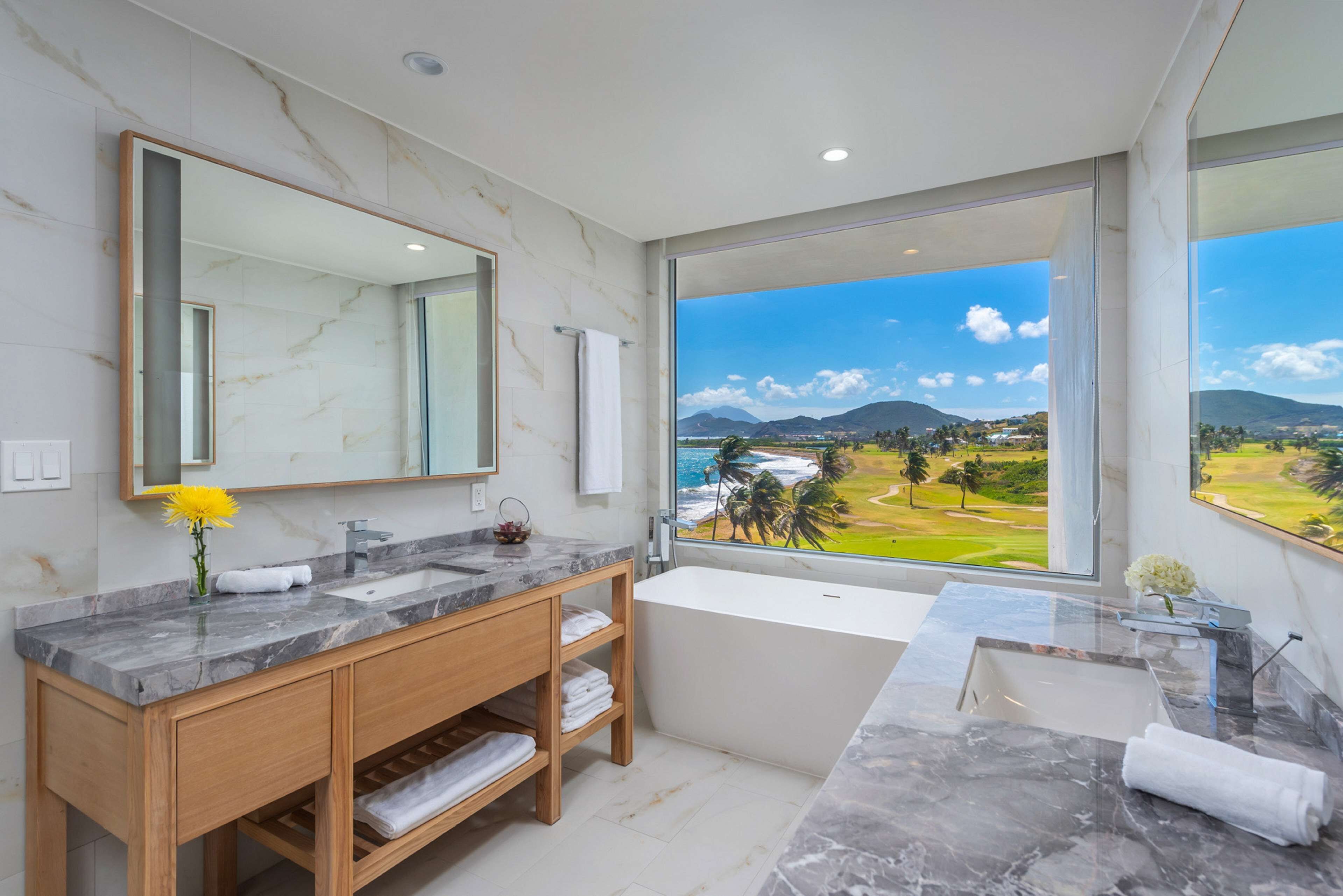 A modern bathroom with a marble vanity, a large mirror, and a bathtub. It features a large window with a view of a golf course and mountains.
