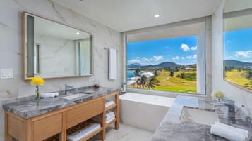 A modern bathroom with a marble vanity, a large mirror, and a bathtub. It features a large window with a view of a golf course and mountains.