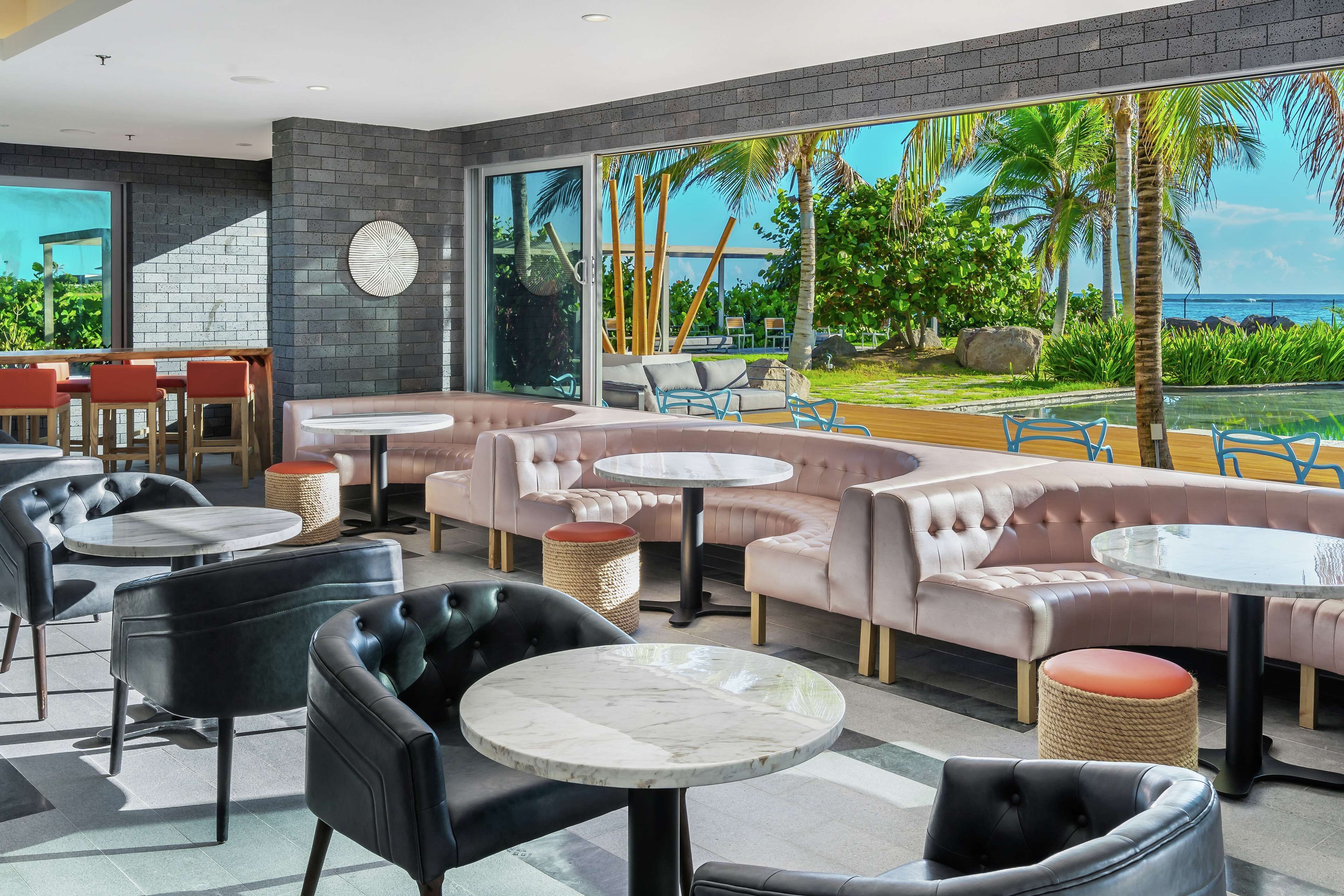 A modern lounge with leather sofas, round tables, and a large window overlooking a pool and tropical scenery.