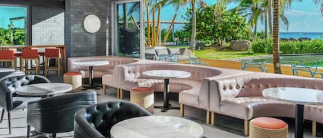 A modern lounge with leather sofas, round tables, and a large window overlooking a pool and tropical scenery.