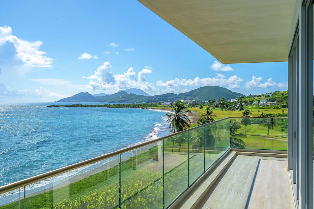 A balcony with a glass railing overlooking a beach, ocean, and distant mountains.