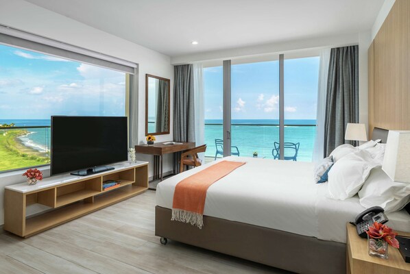 A hotel room with a large bed, a TV on a wooden stand, a desk, and a view of the ocean. - Koi Resort Saint Kitts, Curio Collection by Hilton (Basseterre)
