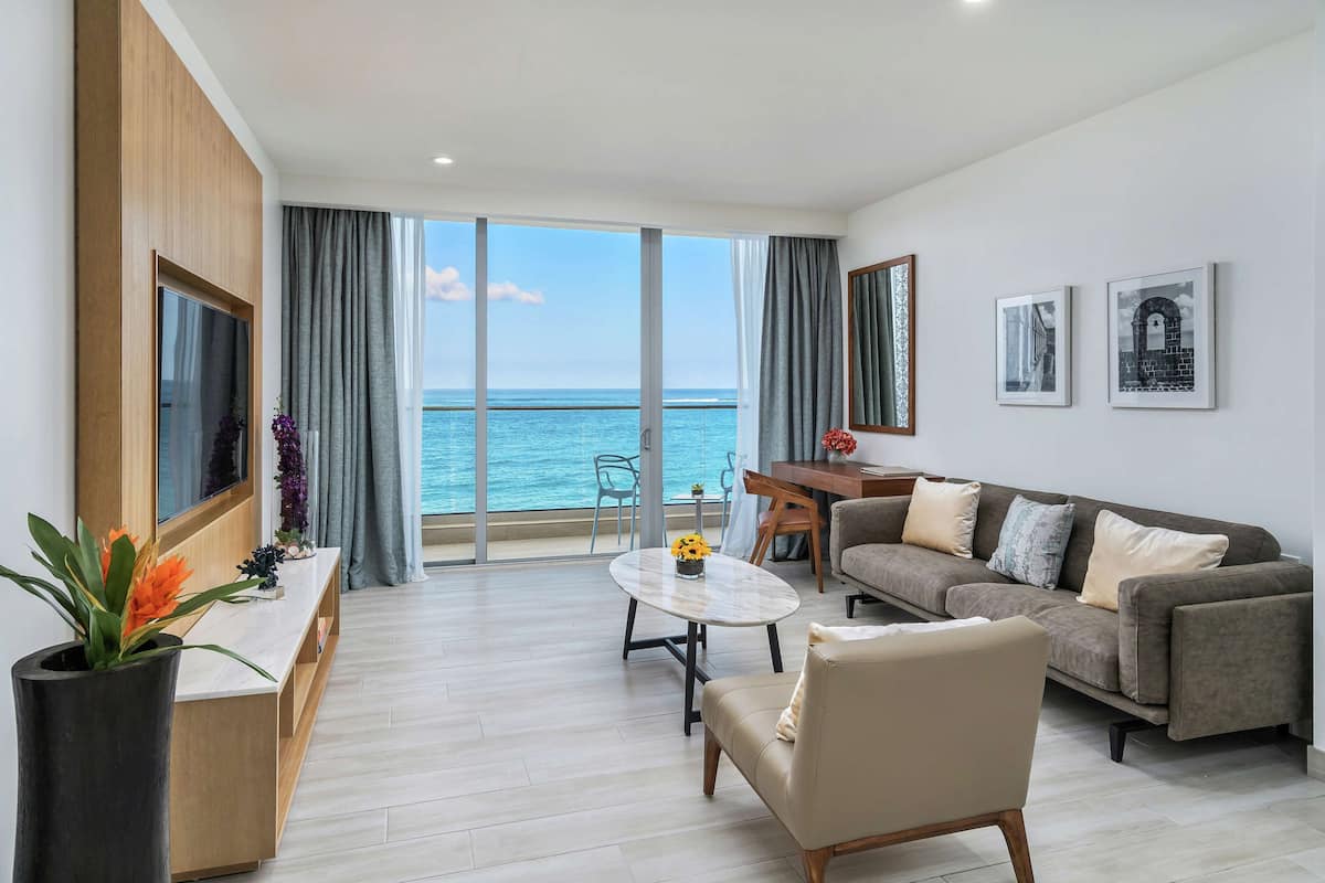 A modern living room with a sofa, coffee table, and a TV. There is a large window with a view of the sea.