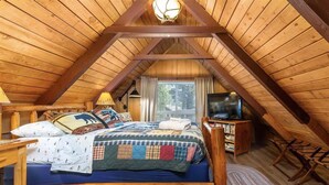 Un dormitorio cálido en el ático con techo de madera, una cama con colcha, un televisor y una pequeña mesa de madera.