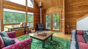 A cozy cabin interior with a wooden interior, a fireplace, large windows, and a green patterned rug. - Trails End | Hot Tub, Starlink, Woodstove (Packwood)