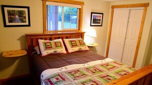 A bedroom with a wooden bed, a window with a view of greenery, a door, and framed pictures on the walls.