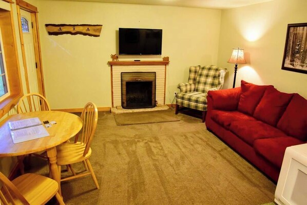 A living room with a fireplace, a television, a plaid armchair, a red sofa, and a dining table with chairs.
