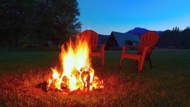 A campfire with two Adirondack chairs in a grassy field with tents in the background.