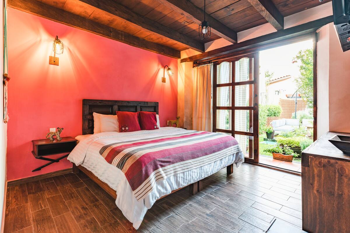 A bedroom with a wooden ceiling, a large bed with a striped blanket, two bedside tables, and a window with curtains.
