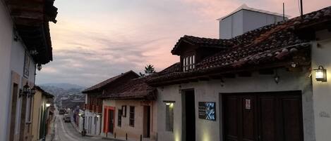 Una calle estrecha bordeada de edificios tradicionales, algunos con tejados de tejas y otros con fachadas planas. Hay farolas y un cartel que indica un hotel. La calle está pavimentada con adoquines.