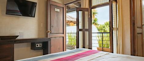 A hotel room with a bed, a desk with a flat-screen TV, a wooden door, and a balcony with plants.