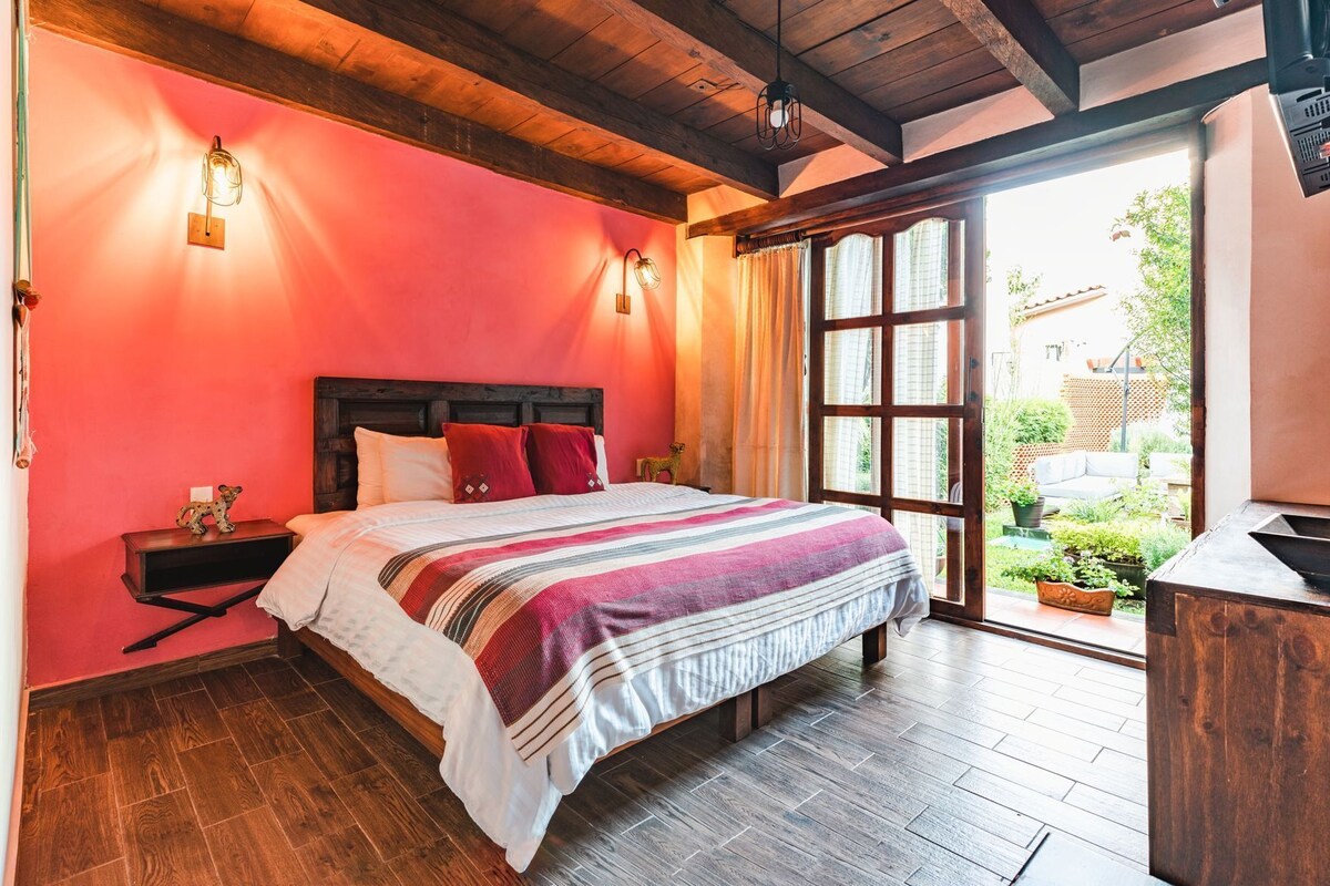 A bedroom with a wooden ceiling, a bed with a striped blanket, a nightstand, and a view of a garden through a window.