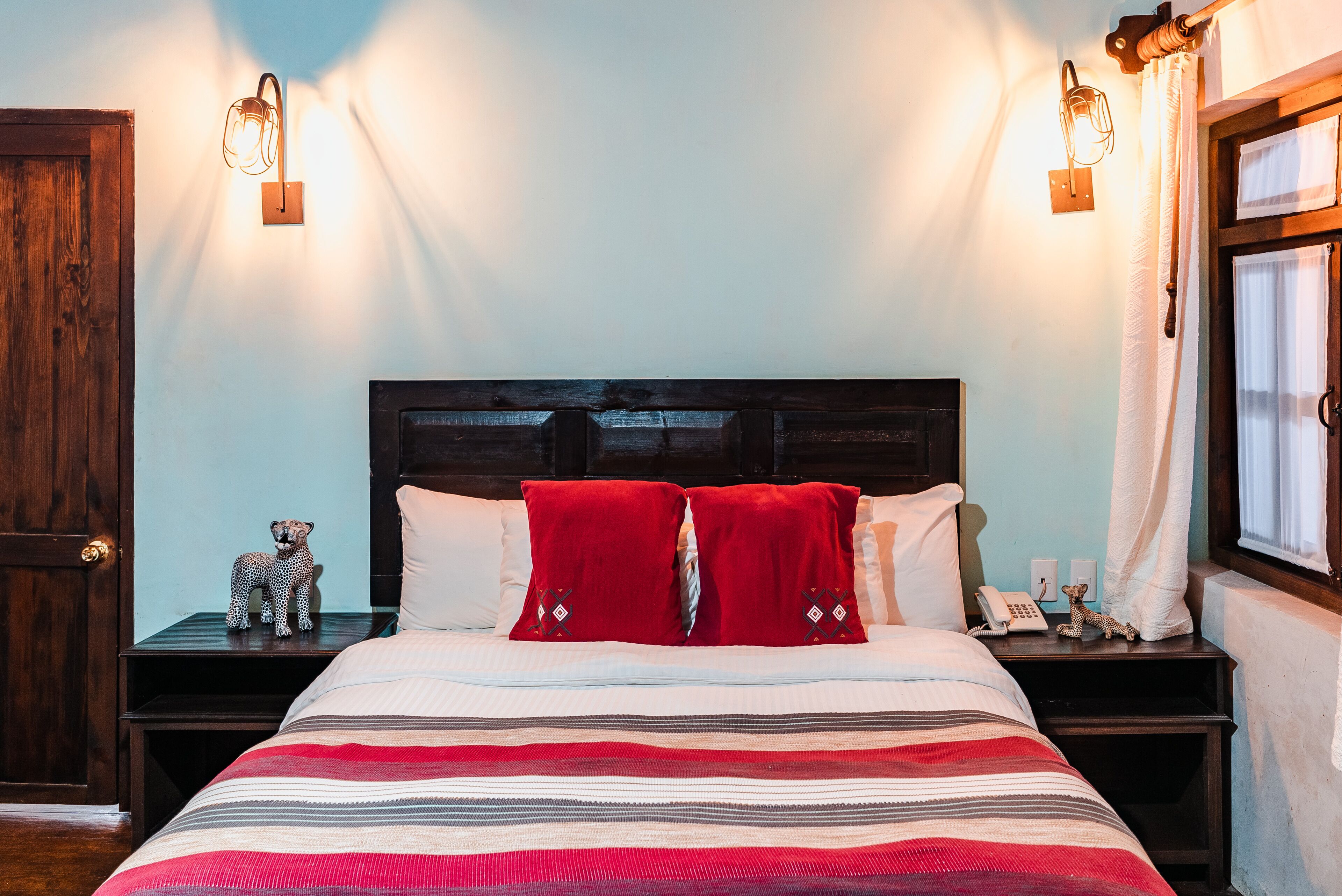 Una cama con ropa de cama a rayas, dos almohadas rojas, una figurita de leopardo y dos lámparas fijadas a la pared.