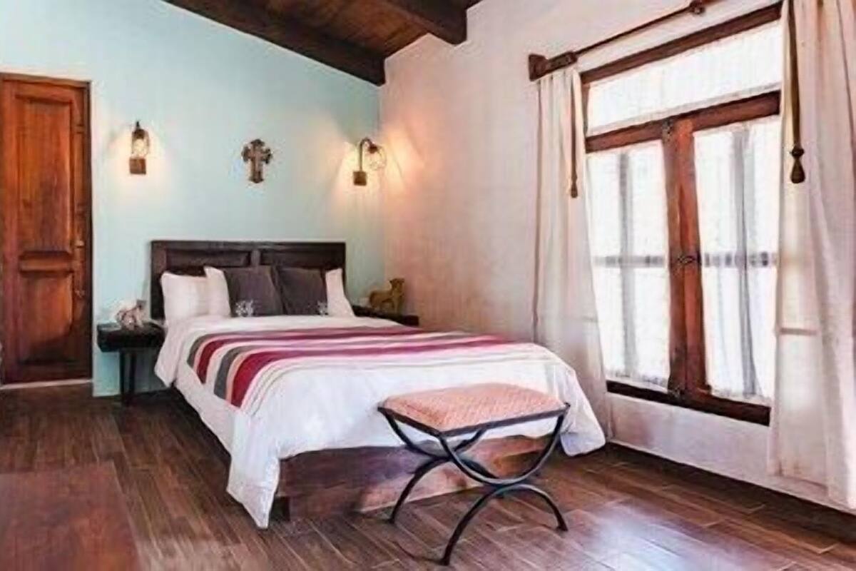 A bedroom with a wooden floor, a bed with a striped bedspread, a nightstand, a wall-mounted light, and a window with curtains.