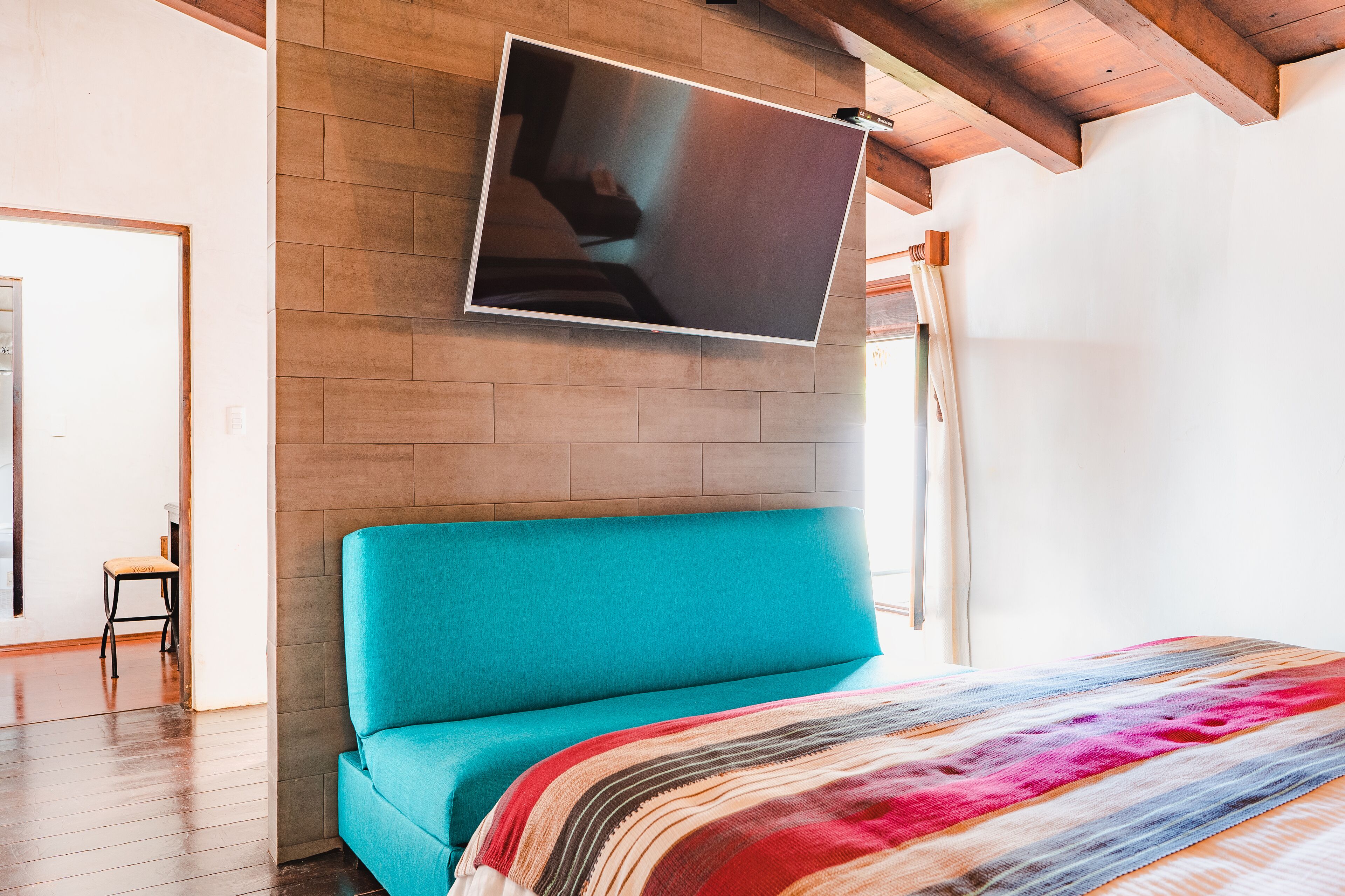A bedroom with a teal bed, a colorful blanket, a mounted TV, and a wooden ceiling.